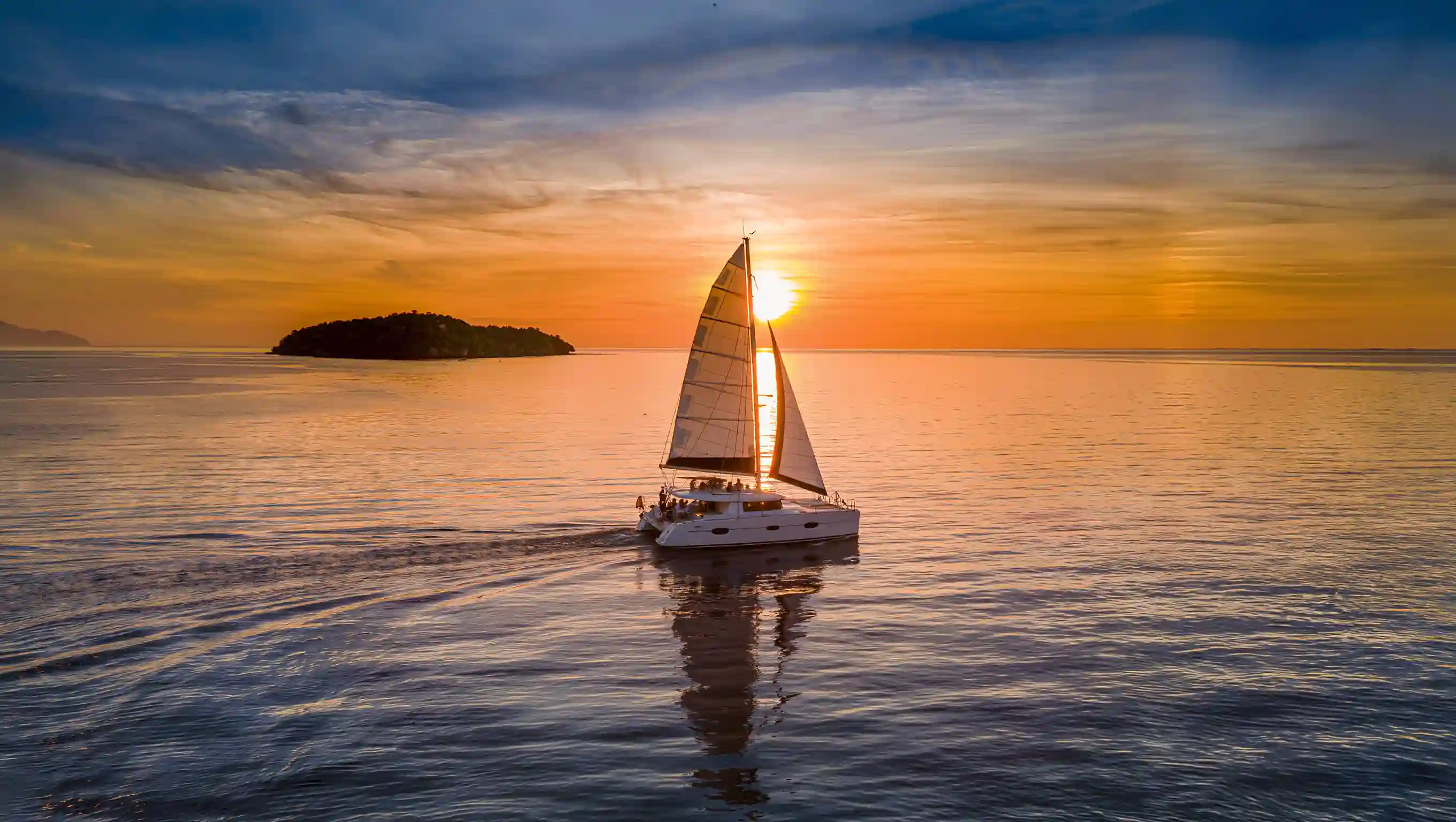 A sailing catamaran under way at sunset, twin hulls and full sail